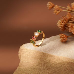 May include: A gold-toned ring featuring four distinct gemstones in a row. The stones include a rectangular olive green gem, a round red gem, a round clear white gem, and a marquise-shaped emerald green gem, all set in a delicate metal band.