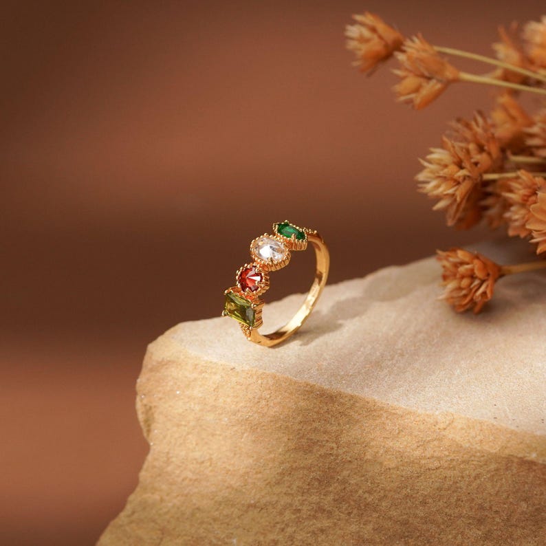May include: A gold-toned ring featuring four distinct gemstones in a row. The stones include a rectangular olive green gem, a round red gem, a round clear white gem, and a marquise-shaped emerald green gem, all set in a delicate metal band.