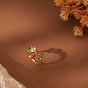 May include: A gold-toned ring featuring four distinct gemstones in a row. The stones include a green oval, a clear oval, a red square, and a green rectangular gem. The band is thin and metallic, showcasing a polished finish with a delicate setting.