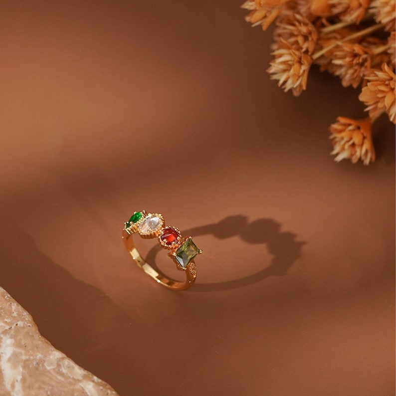 May include: A gold-toned ring featuring four distinct gemstones in a row. The stones include a green oval, a clear oval, a red square, and a green rectangular gem. The band is thin and metallic, showcasing a polished finish with a delicate setting.
