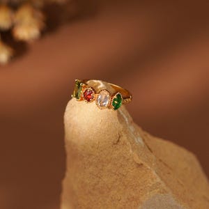 May include: A gold-toned ring featuring four distinct gemstones arranged in a row. The stones include a rectangular olive green gem, a round red gem, a central oval clear white gem, and a marquise-shaped emerald green gem set in metal.