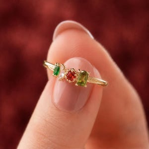 May include: A gold ring featuring four small, multi-colored gemstones arranged in a row. The stones include a small green oval, a clear white stone, a round red stone, and a rectangular green stone, all set in a delicate gold band with metal detailing.