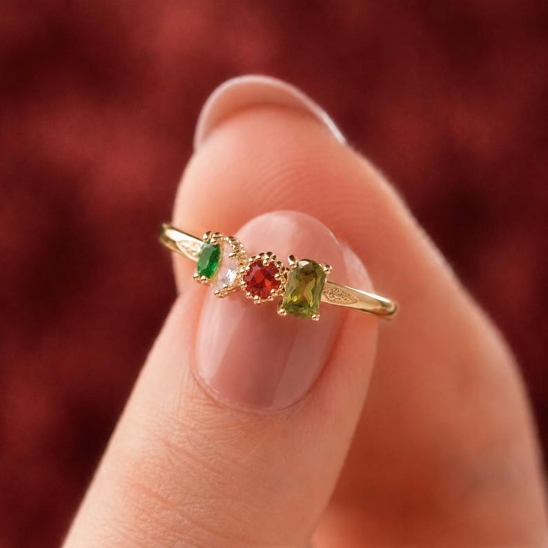 May include: A gold ring featuring four small, multi-colored gemstones arranged in a row. The stones include a small green oval, a clear white stone, a round red stone, and a rectangular green stone, all set in a delicate gold band with metal detailing.