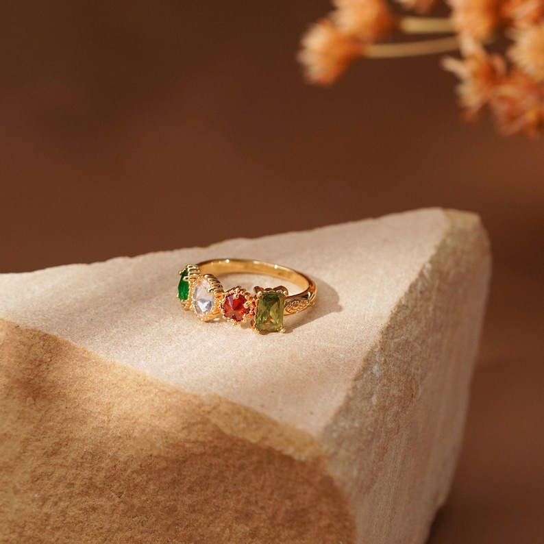 May include: A gold-toned ring featuring four distinct gemstones in a row. The stones include a green pear-cut, a clear marquise-cut, a red round-cut, and a light green rectangular-cut. The metal band has a textured, beaded edge detail.