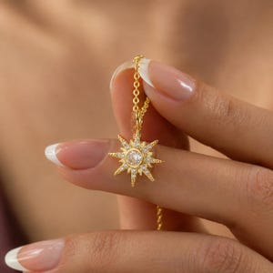 May include: A gold-toned necklace featuring a starburst-shaped pendant. The pendant has a central round clear stone surrounded by smaller clear stones set along the radiating points of the star. The pendant hangs from a delicate gold-toned chain.