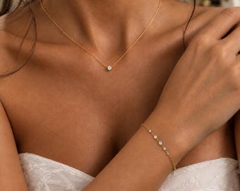 Puede incluir: Un collar de cadena dorada con un colgante de piedra redonda transparente y una pulsera de cadena dorada a juego adornada con cinco pequeñas piedras redondas transparentes espaciadas, sobre una prenda blanca texturizada.