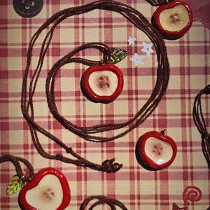 Puede incluir: Cuatro colgantes con forma de manzana en collares de cordón marrón. Cada colgante presenta un borde exterior rojo y un centro color crema con cuatro pequeños agujeros que parecen un botón. Un pequeño dije de hoja metálica está en la parte superior.