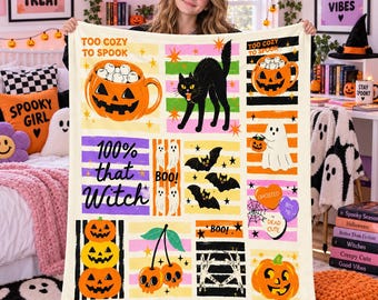 May include: A cream-colored throw blanket featuring a grid of Halloween-themed illustrations including a jack-o'-lantern mug, a black cat, ghosts, bats, and cherries with skull faces. The text reads 'TOO COZY TO SPOOK', '100% that Witch', 'BOO!', and 'Mia'.