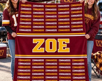 May include: A burgundy fleece blanket featuring the word ZOE in large yellow block letters centered between two horizontal yellow stripes. The top and bottom sections are covered in a repeating pattern of the word COMMANDERS in white.