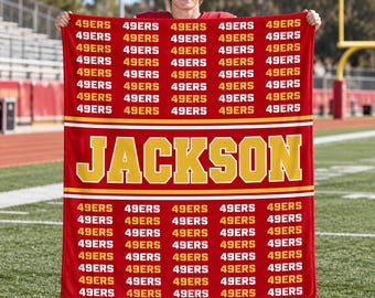 May include: A red rectangular blanket featuring a repeating pattern of the white text 49ERS in horizontal rows. A central horizontal band displays the name JACKSON in large yellow capital letters, bordered by thin white and yellow stripes.