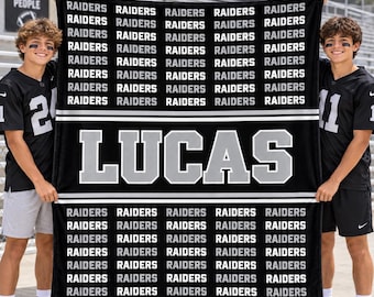 May include: A black and white fleece blanket featuring the repeated word RAIDERS in a grid pattern, with the name LUCAS printed in large, bold, block lettering across the center, framed by two horizontal white stripes.