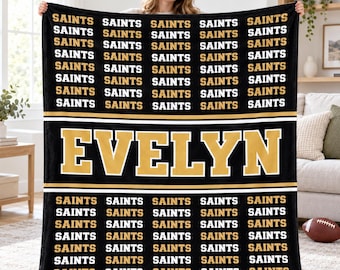 May include: A black rectangular fleece blanket featuring a repeating pattern of the word SAINTS in white and gold text. A central horizontal band displays the name EVELYN in large gold letters with a white outline on a black background.