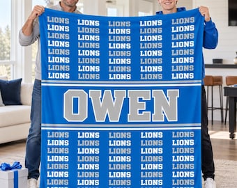 May include: A blue fleece blanket featuring a repeating pattern of the word LIONS in white text, with the name OWEN printed in large white block letters across the center between two horizontal white stripes on a blue background.