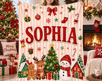 May include: A soft, cream-colored throw blanket with vertical light pink stripes, featuring the name SOPHIA in large red letters. The design includes a central Christmas tree, a snowman, a reindeer, a gingerbread person, and festive holiday icons.