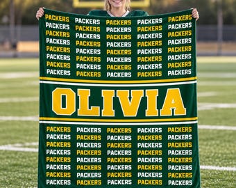 May include: A dark green fleece blanket featuring a repeating pattern of the word PACKERS in white and yellow text, with the name OLIVIA printed in large yellow block letters across a central horizontal band on the front of the fabric.