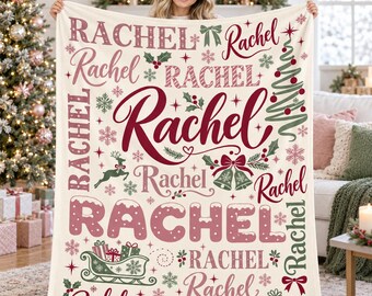 May include: A cream-colored fleece blanket featuring the name Rachel repeated in various red and green holiday-themed fonts. The design includes illustrations of a Christmas tree, a reindeer, bells, snowflakes, holly, and a gift-filled sleigh.