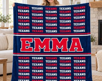 May include: A dark blue fleece blanket featuring a repeating pattern of the word TEXANS in red and white text, with the name EMMA printed in large red letters with a white outline across the center, separated by horizontal white stripes.
