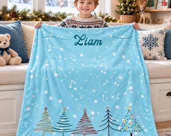 May include: A light blue fleece blanket featuring a white snowflake pattern and the name Liam printed in dark teal script. The bottom edge displays five stylized Christmas tree illustrations in various colors and line designs.
