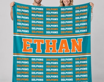 May include: A teal fleece blanket featuring a repeating pattern of the word DOLPHINS in white and orange text. A wide horizontal band across the center displays the name ETHAN in large orange block letters with a white outline.