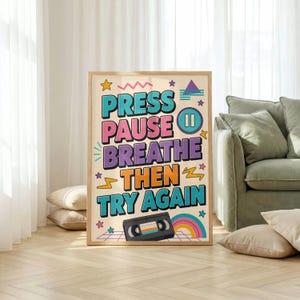 May include: A rectangular wall art print featuring the text PRESS PAUSE BREATHE THEN TRY AGAIN in bold, colorful letters. The design includes a black VHS cassette tape, a pause icon, a rainbow, and various geometric shapes like stars and zigzags.