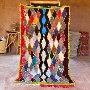 Op de afbeelding: Een kleurrijk, handgemaakt tapijt met een ruitpatroon. Het tapijt heeft een verscheidenheid aan kleuren, waaronder rood, blauw, geel en groen. De randen zijn afgewerkt met een felgroene rand en rode franjes.