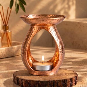 May include: A copper-colored metal oil burner with a hammered texture and an arched base. The burner features a wide, shallow bowl at the top for wax or oil and a central opening at the bottom designed to hold a small, lit tea light candle.