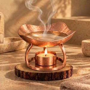 May include: A copper-coloured metal oil burner with a hammered texture, featuring a wide bowl for oil and a lower circular stand for a tea light candle. A lit tea light candle sits in the base, emitting smoke from the bowl above.