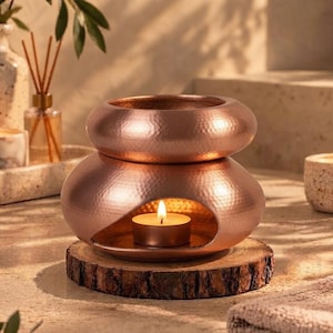 May include: A copper-coloured metal oil burner with a hammered texture, featuring a rounded base with a central opening for a tea light candle and a bowl-shaped top, resting on a circular wooden slice base with a lit candle inside.