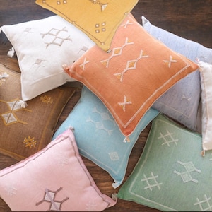 May include: A collection of square throw pillows in various colors, including orange, yellow, pink, and blue. Each pillow features embroidered geometric patterns and decorative tassels. The pillows are arranged on a wooden surface.