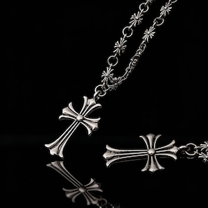 May include: A silver necklace featuring a cross pendant and a separate cross charm. The cross pendant has a detailed design with curved edges and a central stud. The necklace chain has small, star-shaped links. The background is black.