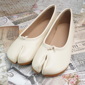 May include: A pair of off-white split-toe ballet flats. The shoes have a rounded toe design with a small bow detail and a separate section for the big toe. The soles are a light brown color.