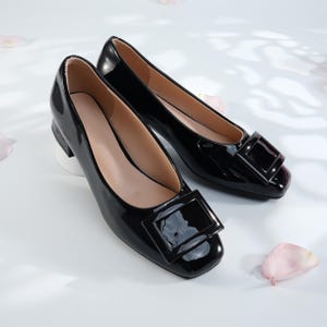 Puede incluir: Un par de bailarinas negras brillantes con hebillas cuadradas. Los zapatos tienen un tacón bajo y una punta redondeada. El interior del zapato es de color beige claro. Los zapatos se exhiben sobre una superficie blanca con pétalos de flores rosas.