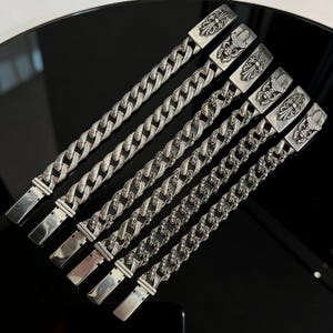 May include: Six silver-toned chain bracelets with ornate rectangular clasps. The bracelets feature a curb chain design, with each link interlocked. The clasps are decorated with floral and shield-like motifs. The bracelets are displayed on a reflective black surface.