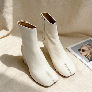 Può includere: Un paio di stivaletti alla caviglia in pelle bianco sporco con design a punta divisa. Gli stivaletti hanno un tacco arrotondato e un gambale alto, con dettagli di cucitura visibili. La fodera interna è di colore marrone chiaro. Una rivista con la parola "NINE" è visibile sullo sfondo.