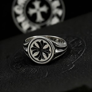 Könnte beinhalten: Ein silberner Ring mit einem kreisförmigen Gesicht mit einem stilisierten Kreuzdesign. Der Ring hat ein detailliertes, strukturiertes Band und einen dunklen Hintergrund. Ein ähnliches Design ist im Hintergrund sichtbar und erzeugt einen geschichteten Effekt. Der Ring ist ein Schmuckstück.