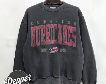 Puede incluir: Sudadera gris oscuro con el texto "CAROLINA HURRICANES" en rojo, con "EST. 1972" debajo. La sudadera está colgada de una percha de madera.
