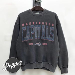 Op de afbeelding: Een donkergrijze sweatshirt met de tekst "WASHINGTON CAPITALS" in grote, vervaagde letters over de borst. Daaronder staat "EST 1974". De sweatshirt hangt aan een houten kleerhanger.