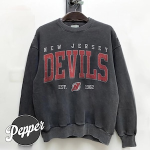 Może przedstawiać: Szara bluza z napisem "NEW JERSEY DEVILS" w czerwonych literach. Pod słowem "DEVILS" znajduje się napis "EST. 1982" i logo. Bluza wisi na drewnianym wieszaku.