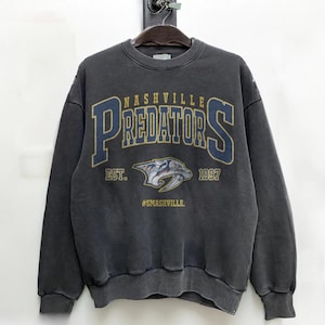 May include: Dark gray crewneck sweatshirt featuring the text "NASHVILLE PREDATORS" in navy blue and gold lettering. Below the text is a logo of a saber-toothed tiger and the text "EST. 1997" and "#SMASHVILLE."