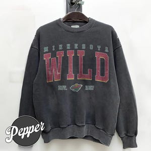 Op de afbeelding: Donkergrijze sweatshirt met de tekst "MINNESOTA WILD" in bordeauxrode en gouden letters. De sweatshirt heeft ook een logo en de tekst "EST. 1997". De sweatshirt hangt aan een houten hanger.