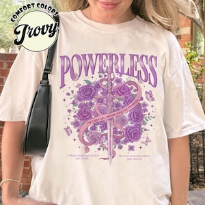 Op de afbeelding: Crèmekleurig T-shirt met het woord "POWERLESS" in paars, met een zwaardontwerp omringd door paarse rozen en vlinders. Het shirt bevat de tekst "FOR EVERY GIRL WHO HAS EVER FELT POWERLESS."