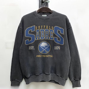 May include: Dark gray sweatshirt with the text "BUFFALO SABRES" in blue and gold lettering. The sweatshirt also features a logo with a buffalo and crossed hockey sticks, and the text "EST. 1970" and "#BUILT FOR BATTLE."