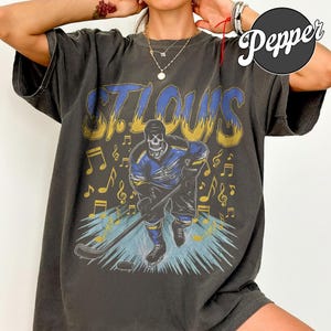 Puede incluir: Camiseta gris oscuro con un esqueleto jugando al hockey y el texto "ST. LOUIS" en azul y dorado. El diseño incluye notas musicales y un palo de hockey. La camiseta tiene un aspecto vintage y desgastado.