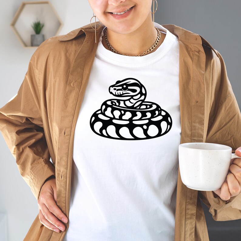 May include: White t-shirt with a black and white illustration of a coiled snake. The snake design is in the centre of the shirt. The person in the image is wearing a brown jacket and holding a white mug.