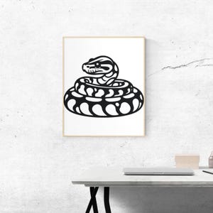 May include: A framed art print showing a coiled snake illustration in black and white. The snake has detailed patterns and a focused gaze, set against a white background. The artwork is displayed in a gold-coloured frame.