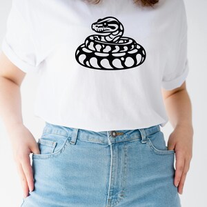 May include: White t-shirt with a black and white coiled snake graphic. The snake design has a detailed head and body. The shirt is made of a soft material, suitable for casual wear. The t-shirt is a versatile item.