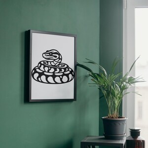 May include: A framed black and white print of a coiled snake. The snake illustration features a detailed head and a patterned body. The artwork is displayed on a green wall, with a potted plant nearby.