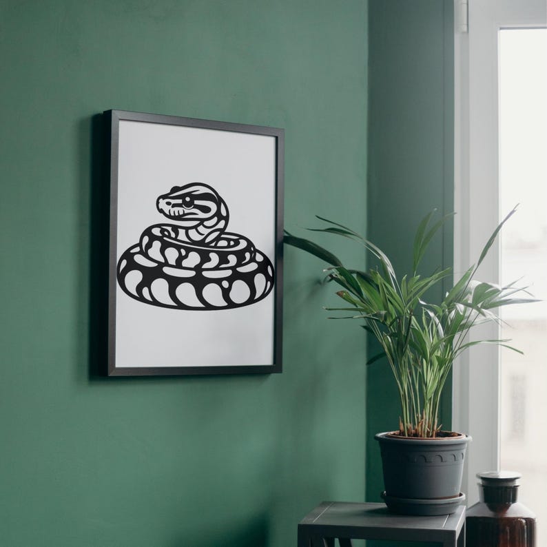 May include: A framed black and white print of a coiled snake. The snake illustration features a detailed head and a patterned body. The artwork is displayed on a green wall, with a potted plant nearby.