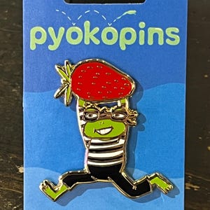 May include: A colorful enamel pin featuring a cartoon frog wearing glasses and a striped shirt, with a strawberry for a hat. The pin is on a blue backing card with the word "pyokopins" in green letters.