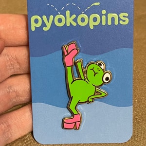 May include: A gold-colored enamel pin featuring a green frog wearing pink high heels. The frog is standing on one leg with its other leg raised in the air. The pin is on a blue and white background with the text "pyokopins" at the top.